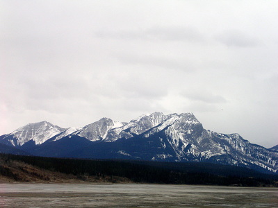 [The lake area just looks like ground with some frozen water spots. There are small hillsides at the lake's edge, but the Rocky Mountains in the distance tower above them. Lower levels appear to be evergreens while the rest is snow-dusted.]